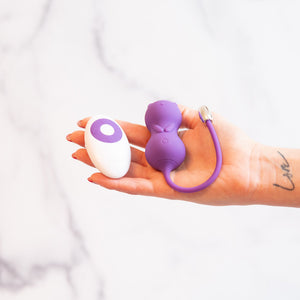 Kitty Kegel Ball Vibrator  has a cat shape in the color purple with a long silicone strap sitting in a white model's hand with a white and purple remote control to the left.