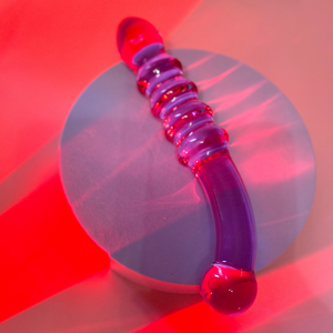 Translucent purple glass dildo. One half of the shaft has five textured ribs tracing down to a cone-shaped tip, and the other side of the shaft is smooth, extending down to a bulbous, rounded tip.